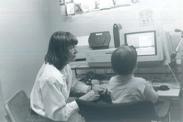 Alle kouluikäinen lapsi pelaa tietokoneella peliä, nainen istuu vieressä ohjaamassa.