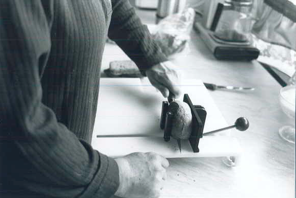 Ihminen leikkaa leipää veitsellä yhdellä kädellä, leipä on kiinni apuvälineen avulla leikkuulaudassa.